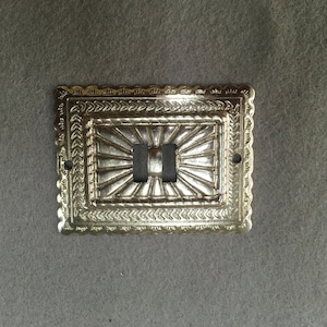 May include: A gold-colored metal light switch cover with an ornate design. The cover has a decorative pattern of radiating lines and a border of small triangles.