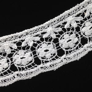 May include: White crocheted lace trim featuring a delicate floral design. The lace has a scalloped edge and is ideal for adding a touch of elegance to any project.