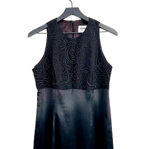 May include: A sleeveless black dress hanging on a black hanger. The bodice features an intricate, raised, swirling design. The skirt is a smooth, satin-like material. The dress is a formal or semi-formal garment.