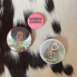 May include: Three round buttons with unique designs. One pink button reads "WEIRDNESS IS UPON US." Another features a portrait with a green and red collar. The third button shows an oyster and the word "Suffering."