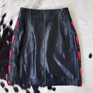May include: A black leather skirt featuring vertical white contrast stitching and decorative red teardrop-shaped cutouts along both side panels. The skirt has a high-waisted design with a straight silhouette and a smooth, polished finish.