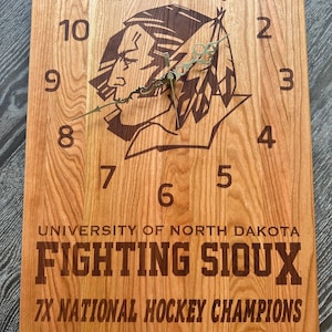 May include: A wooden clock with a brown and black image of a Native American man with feathers. The clock face has Roman numerals and the text "UNIVERSITY OF NORTH DAKOTA FIGHTING SIOUX 7X NATIONAL HOCKEY CHAMPIONS 1959-1963-1980-1982-1987-1997-2000".