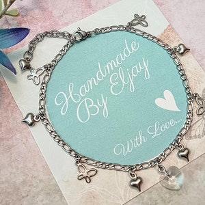 May include: A silver chain bracelet with multiple charms, including hearts and butterflies. The bracelet is on a light blue background with the text "Handmade By Eljay With Love..."
