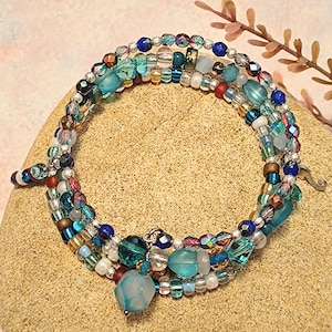 May include: A multi-strand beaded bracelet with blue, teal, and iridescent beads. The bracelet has round, faceted, and irregular beads. A silver clasp secures the bracelet. The bracelet is approximately 18 cm in circumference.