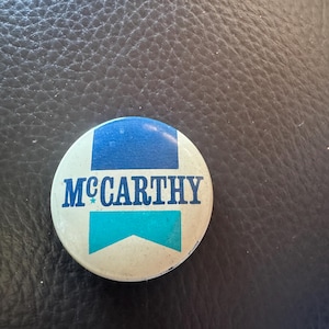May include: A round, vintage button with the name "McCarthy" in navy blue lettering. The button has a white background with blue and teal accents. The button is slightly worn, suggesting age.