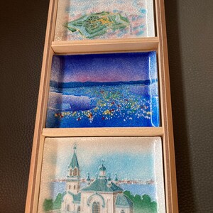 May include: A wooden box with three small decorative plates inside. The plates are painted with scenes of a city, a church, and a landscape. The plates are in a wooden box with a light brown finish.