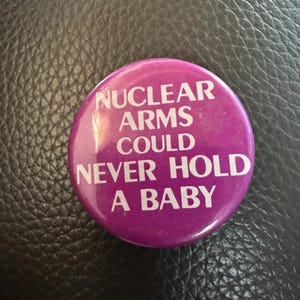 May include: A round, purple button with white text that reads "NUCLEAR ARMS COULD NEVER HOLD A BABY". The button is set against a dark, textured background.