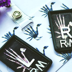 Pode incluir: Duas etiquetas retangulares pretas com um desenho de mão esquelética branca e as letras "RM" e "L" em um fundo padronizado. Um retrator de crachá roxo e um porta-crachá transparente também são visíveis.