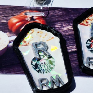 Può includere: Due porta badge a tema Starbucks con bordi neri, con il logo Starbucks e le lettere "R", "L" e "RM". Lo sfondo include una tazza di caffè con un design a cuore e una zucca.