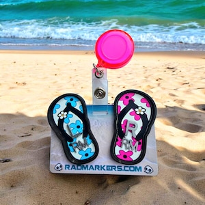 May include: A beach-themed badge reel with two flip-flop designs. One flip-flop is blue with white and light blue flowers, the other is pink with white and pink flowers. The reel has a pink disc and the text "RADMARKERS.COM".