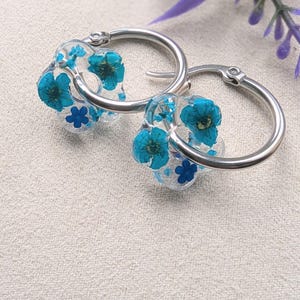 May include: A pair of silver hoop earrings with a clear resin charm featuring pressed blue flowers.