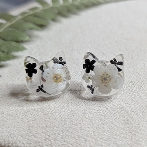 May include: A pair of clear resin cat-shaped earrings with white and black pressed flowers inside.