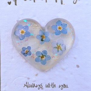 May include: A heart-shaped Pocket Hug featuring blue forget-me-not flowers set in clear resin. The card reads "Pocket Hug" and "Always with you". An Etsy QR code is also present.