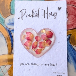 May include: A white card with the text "Pocket Hug" and a heart-shaped resin piece with pink flower petals and gold flakes. The text "You are always in my heart" is written below the resin piece.