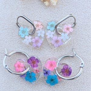 May include: Four pairs of earrings with floral designs. Two pairs feature heart-shaped pendants with colorful pressed flowers and silver hoops. The other two pairs have clear, round pendants with pressed flowers and silver hoops.