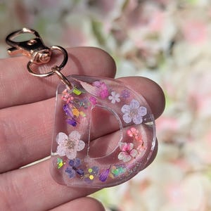 May include: A clear resin keychain in the shape of the letter D, embedded with small dried pink and purple flowers, green foliage, and iridescent glitter flakes. The letter is attached to a rose gold-colored metal lobster clasp hardware.