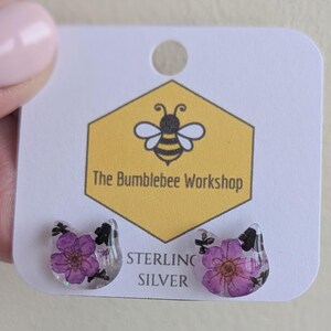 May include: A pair of sterling silver cat-shaped stud earrings. Each earring features a clear resin design with a purple flower and black accents. The earrings are displayed on a white card with the text "The Bumblebee Workshop".