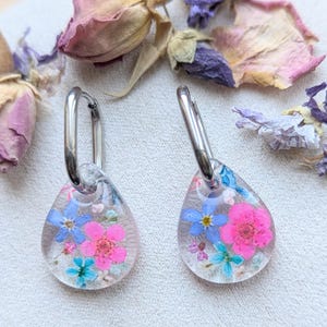 May include: A pair of silver hoop earrings with teardrop-shaped resin charms. The charms are filled with pressed flowers in shades of pink, blue, and green.