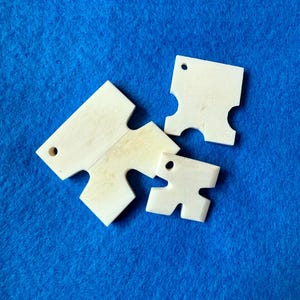 Peut inclure: Trois pièces de puzzle blanc cassé, aux bords arrondis et petits trous, disposées sur une surface en feutre bleu. Les pièces varient en taille, avec une pièce plus grande au centre et deux plus petites. Les formes ont un design unique et emboîtant.