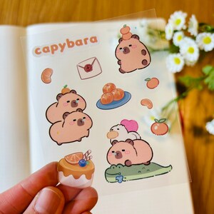 Pu&ograve; includere: Un foglio di adesivi trasparenti con illustrazioni di capibara con fette d'arancia, una lettera e una torta. Gli adesivi sono etichettati "capybara".