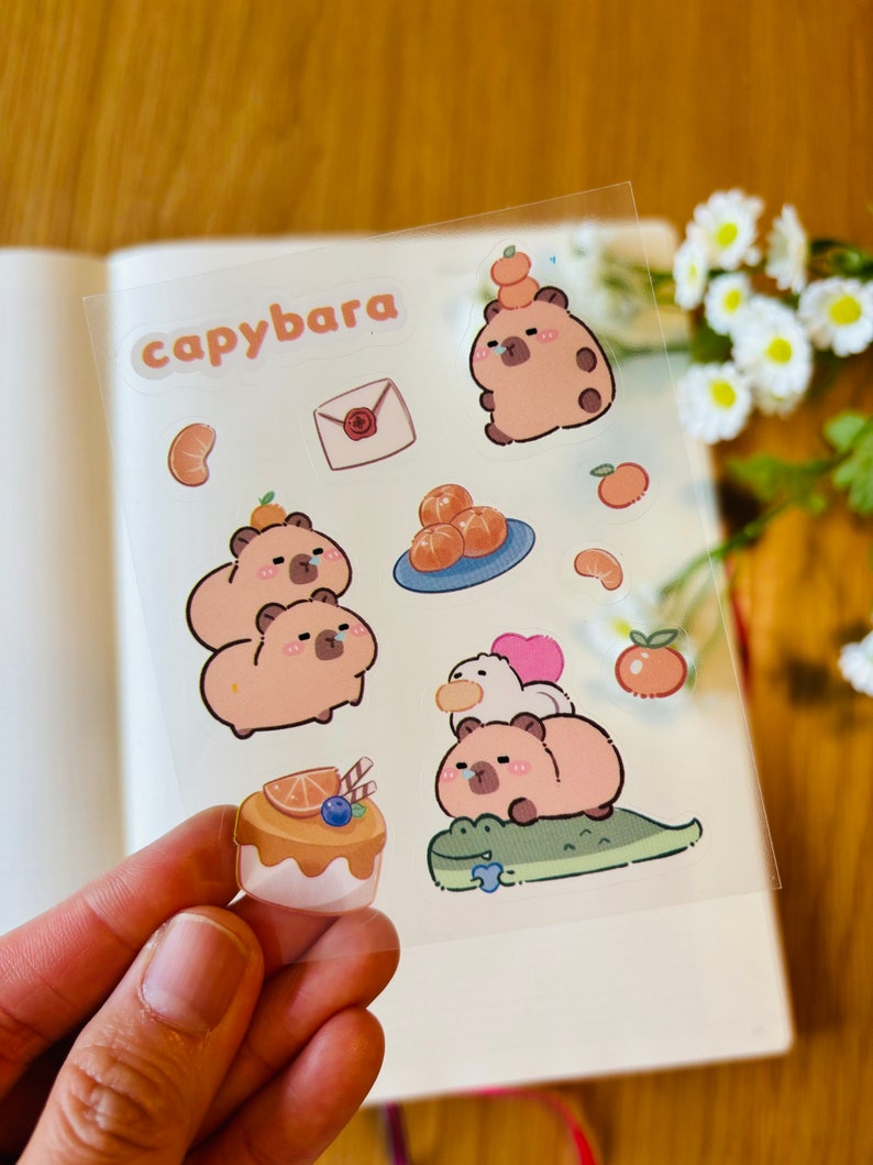 Pu&ograve; includere: Un foglio di adesivi trasparenti con illustrazioni di capibara con fette d'arancia, una lettera e una torta. Gli adesivi sono etichettati "capybara".