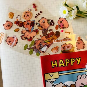 Pu&ograve; includere: Un foglio di adesivi trasparenti con simpatici capibara dei cartoni animati in varie pose. Gli adesivi sono su uno sfondo bianco a griglia. Gli adesivi includono le parole "cute", "nice" e "capybara".