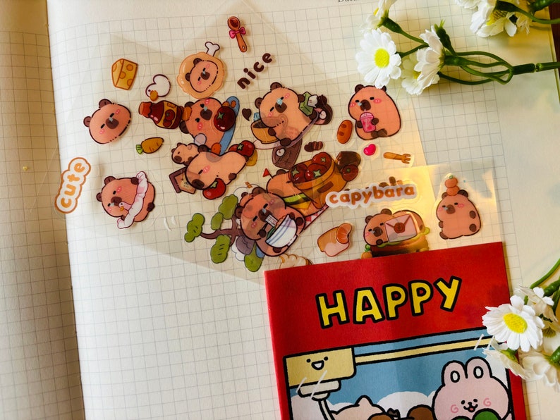 Pu&ograve; includere: Un foglio di adesivi trasparenti con simpatici capibara dei cartoni animati in varie pose. Gli adesivi sono su uno sfondo bianco a griglia. Gli adesivi includono le parole "cute", "nice" e "capybara".