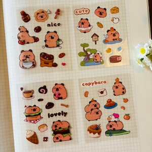 Pu&ograve; includere: Un foglio di adesivi trasparenti con simpatici capibara dei cartoni animati in varie pose e ambientazioni. Gli adesivi includono frasi come "cute", "nice", "lovely" e "capybara".