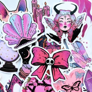 May include: A collection of stickers featuring a variety of designs in shades of pink, purple, and black. The stickers include a seashell, candles, a horned woman with wings, a bow with a skull, crystals, and a butterfly.