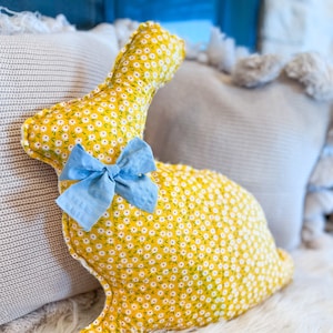 May include: A yellow bunny-shaped pillow with a white floral pattern and a blue bow. The pillow is sitting on a white fluffy surface.