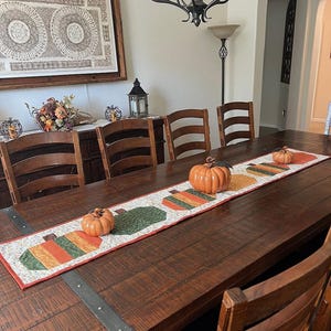 Pode incluir: Uma mesa de jantar de madeira decorada para uma celebra&ccedil;&atilde;o de outono. Uma toalha de mesa decorativa com desenhos de ab&oacute;boras e ab&oacute;boras de cer&acirc;mica s&atilde;o exibidas. A mesa &eacute; cercada por cadeiras de madeira.