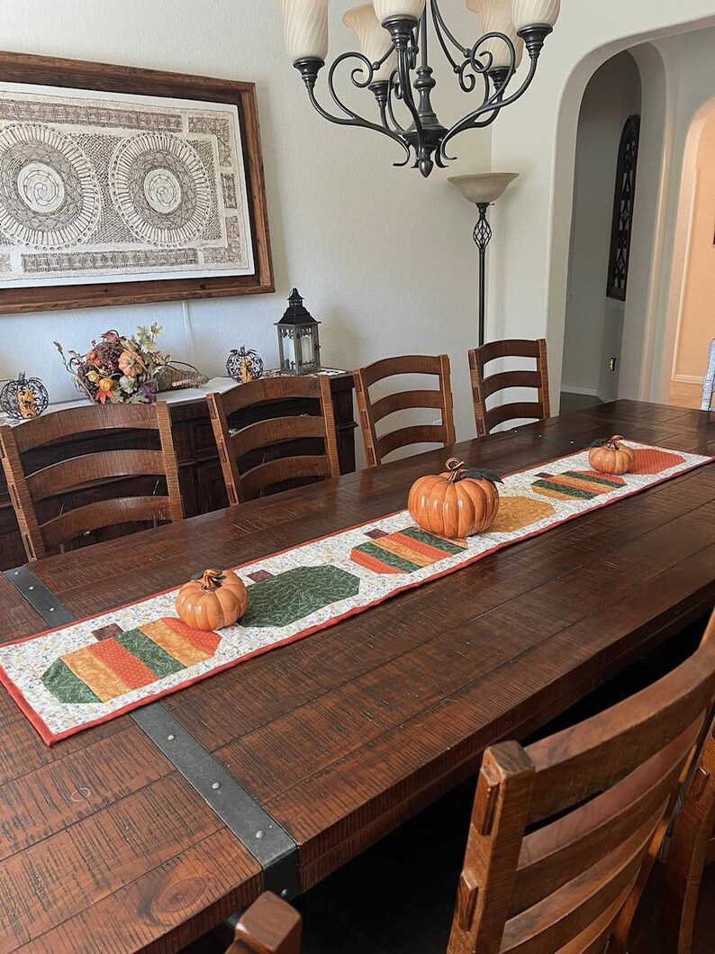 Pode incluir: Uma mesa de jantar de madeira decorada para uma celebra&ccedil;&atilde;o de outono. Uma toalha de mesa decorativa com desenhos de ab&oacute;boras e ab&oacute;boras de cer&acirc;mica s&atilde;o exibidas. A mesa &eacute; cercada por cadeiras de madeira.