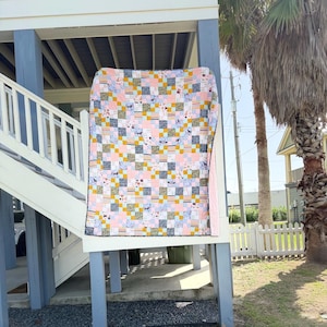 May include: A patchwork quilt featuring a grid of squares in various patterns and colors, including light pink, mustard yellow, white, and dark gray. The fabric squares display small illustrations of dogs, stripes, and geometric shapes throughout.