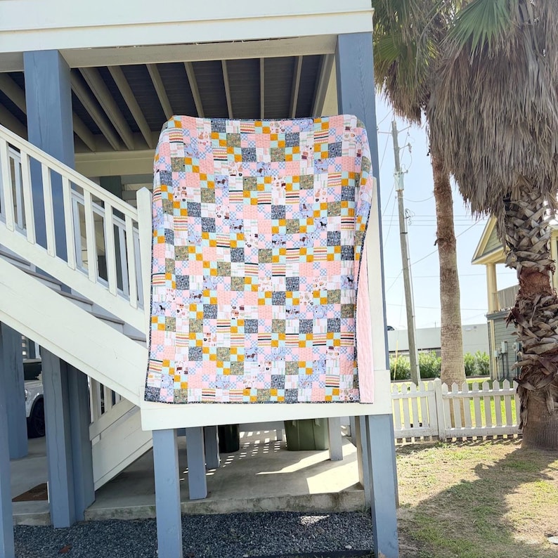 May include: A patchwork quilt featuring a grid of squares in various patterns and colors, including light pink, mustard yellow, white, and dark gray. The fabric squares display small illustrations of dogs, stripes, and geometric shapes throughout.