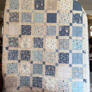 Może przedstawiać: Duża, ręcznie robiona kołdra z patchworkowym wzorem. Kołdra ma kwadraty w różnych odcieniach niebieskiego, beżowego i białego, ze wzorami muszli, rozgwiazd i kształtów geometrycznych. Kołdra jest prawdopodobnie wykonana z bawełny.