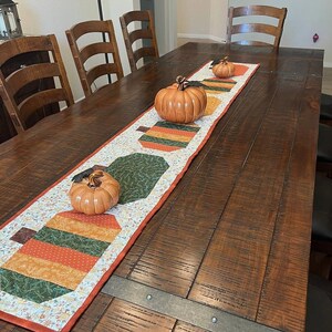 Pode incluir: Uma mesa de jantar de madeira com um caminho de mesa decorativo com tema de outono e tr&ecirc;s ab&oacute;boras de cer&acirc;mica. O caminho de mesa apresenta um design de patchwork com formas de ab&oacute;bora laranja, verde e castanha. As ab&oacute;boras s&atilde;o de v&aacute;rios tamanhos e tons de laranja.