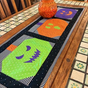 Pode incluir: Um caminho de mesa com tema de Halloween com ab&oacute;boras aplicadas em laranja, roxo e verde. O caminho de mesa tem uma borda preta com bolinhas brancas e listras pretas e brancas. Um pote de vidro laranja cheio de doces laranjas est&aacute; em cima.