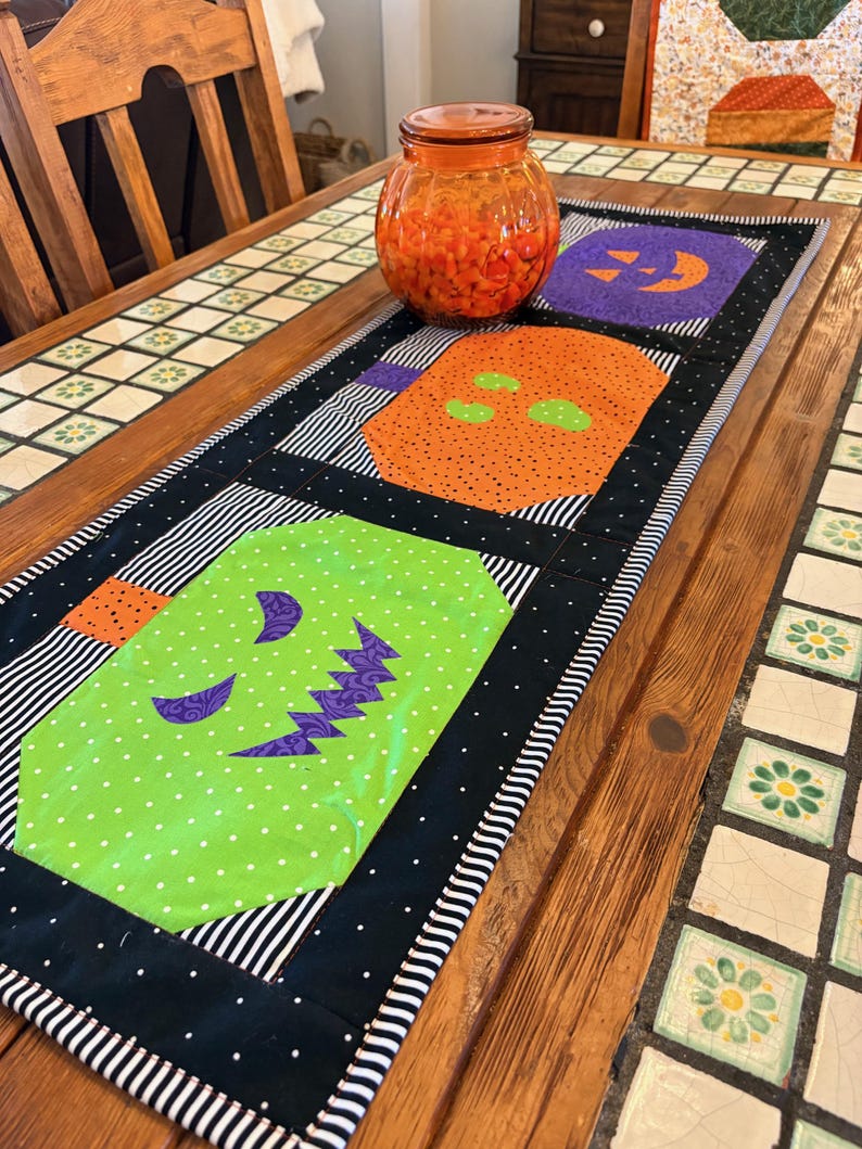 Pode incluir: Um caminho de mesa com tema de Halloween com ab&oacute;boras aplicadas em laranja, roxo e verde. O caminho de mesa tem uma borda preta com bolinhas brancas e listras pretas e brancas. Um pote de vidro laranja cheio de doces laranjas est&aacute; em cima.