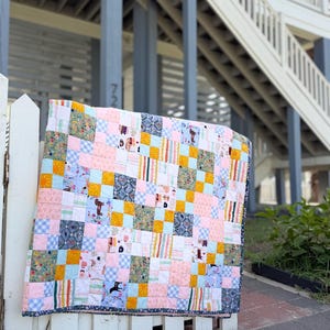 May include: A patchwork quilt featuring a grid of squares in various patterns and colors, including light pink, mustard yellow, light blue gingham, and floral prints. The quilt has a dark blue border with a small white floral pattern throughout.