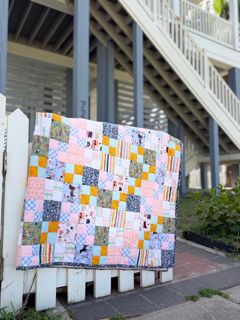 May include: A patchwork quilt featuring a grid of squares in various patterns and colors, including light pink, mustard yellow, light blue gingham, and floral prints. The quilt has a dark blue border with a small white floral pattern throughout.