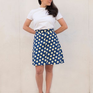 May include: A woman wearing a white t-shirt and a blue skirt with a white and yellow floral pattern. She is standing in front of a white wall and wearing black heeled sandals.