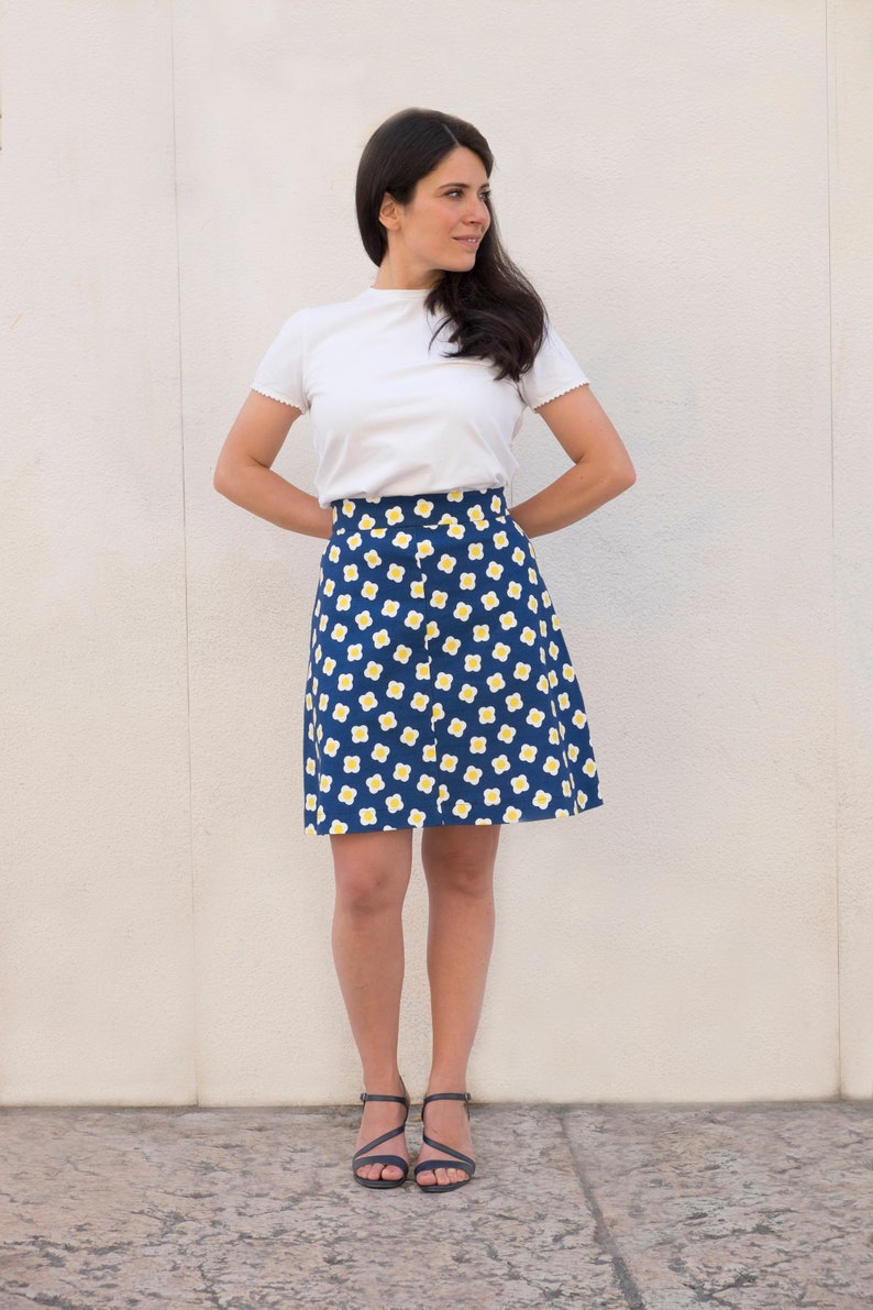 May include: A woman wearing a white t-shirt and a blue skirt with a white and yellow floral pattern. She is standing in front of a white wall and wearing black heeled sandals.