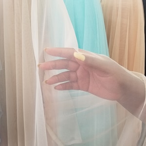 May include: A hand holding a piece of white tulle fabric. The fabric is sheer and has a delicate texture. The fabric is layered over a turquoise blue tulle fabric and a beige tulle fabric.