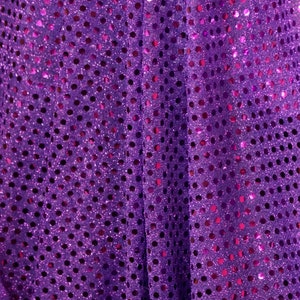 May include: Close-up of a purple fabric with a sequined design. The fabric has a shiny, glittery texture with small, round sequins scattered across the surface. The sequins are a darker shade of purple, creating a contrast against the lighter background.