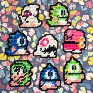 Peut inclure: Une collection d'écussons de personnages pixelisés colorés. Les écussons présentent diverses créatures dans des tons de blanc, vert, rose, bleu et noir. Les écussons sont disposés sur un fond à motifs avec des papillons et des motifs floraux.