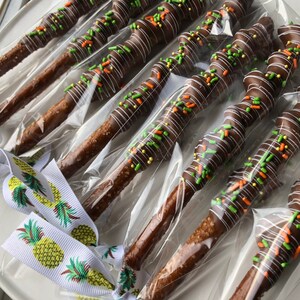May include: A close-up of a dozen chocolate-covered pretzel sticks, each individually wrapped in clear plastic. The pretzels are decorated with white chocolate drizzle, green and orange sprinkles, and yellow sprinkles. The pretzels are tied with a white ribbon with a pineapple pattern.