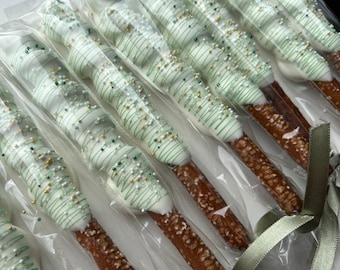 May include: A close-up of a group of pretzel rods dipped in white chocolate and decorated with green and gold sprinkles. The pretzels are individually wrapped in clear plastic bags and tied with a green ribbon.