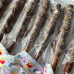 May include: A group of chocolate-covered pretzel rods, each individually wrapped in clear plastic. The pretzels are decorated with white chocolate drizzle and colorful sprinkles. A Winnie the Pooh themed ribbon is tied around the bundle.
