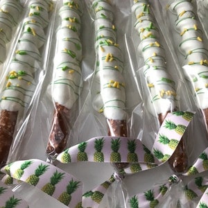 May include: A close-up of several pretzel rods dipped in white chocolate and decorated with green and yellow sprinkles. The pretzels are individually wrapped in clear plastic bags and tied with a pink and green ribbon with a pineapple print.