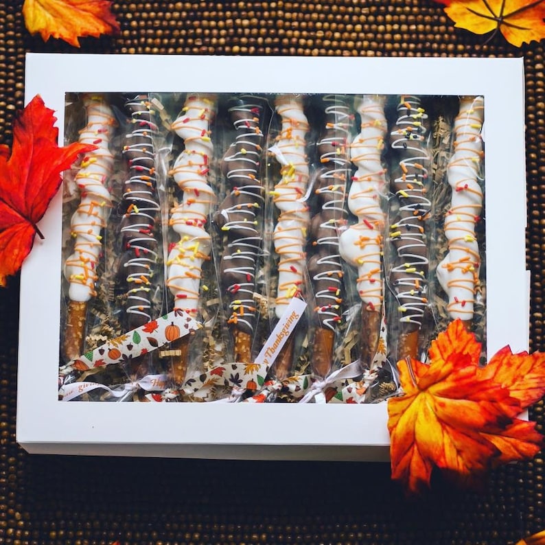May include: A white box filled with chocolate-covered pretzel sticks decorated with white and orange chocolate drizzle and sprinkles. The box is tied with a ribbon and has a "Happy Thanksgiving" tag.